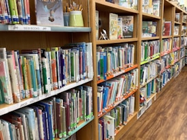 A library shelf filled with colorful children's books organized neatly. A variety of books with vivid covers and titles are visible, alongside some upright display books and a holder with pencils.