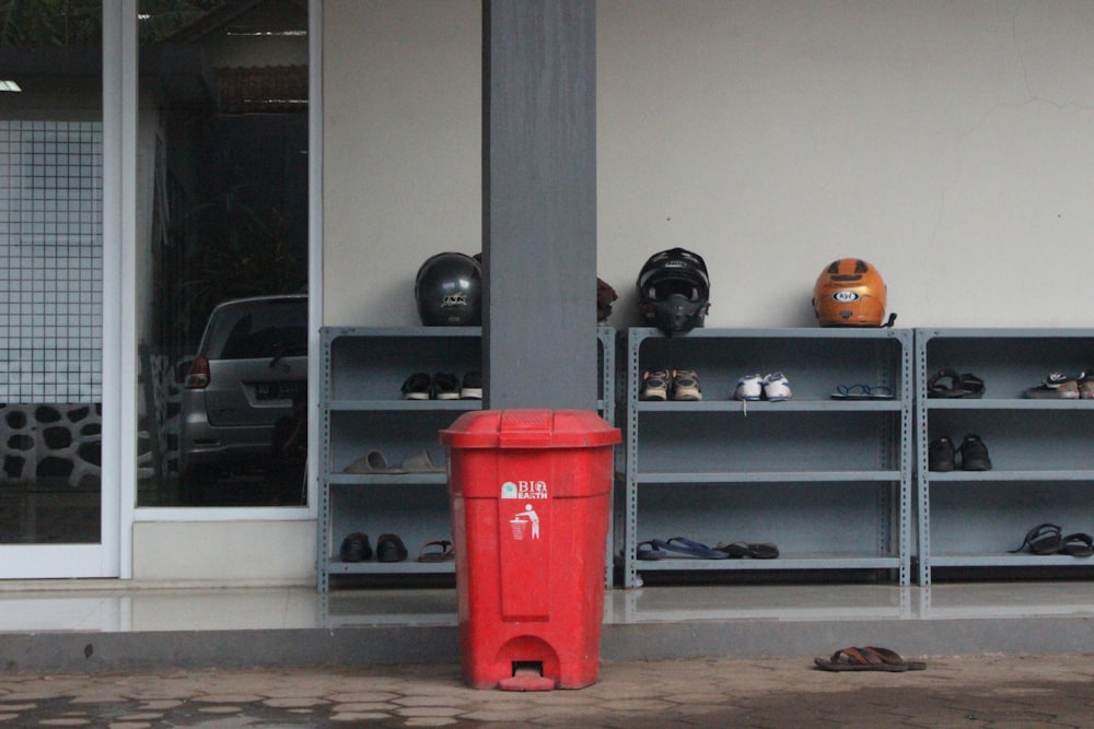 a red garbage can next to a shelf with objects on it