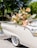 A vintage cream-colored car adorned with an abundant and colorful floral arrangement, including roses and anthuriums. The car is parked outdoors on a sunny day, with green trees in the background.