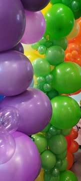 A collection of vibrant, colorful balloons featuring various sizes and colors, including purple, green, orange, and hints of yellow. The balloons create a festive and lively atmosphere with their glossy surfaces reflecting light.
