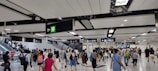 A busy indoor environment with numerous people walking in various directions, indicative of a transportation hub. Overhead signs with directions and exit indicators are visible, along with escalators carrying people to different levels. The ceiling is modern with lighting panels.