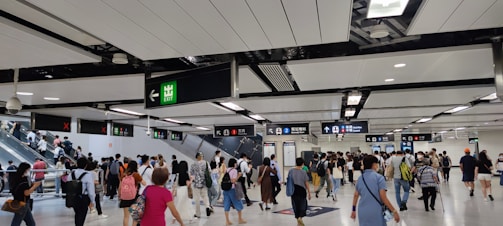 A busy indoor environment with numerous people walking in various directions, indicative of a transportation hub. Overhead signs with directions and exit indicators are visible, along with escalators carrying people to different levels. The ceiling is modern with lighting panels.