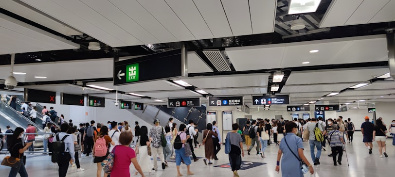 A busy indoor environment with numerous people walking in various directions, indicative of a transportation hub. Overhead signs with directions and exit indicators are visible, along with escalators carrying people to different levels. The ceiling is modern with lighting panels.