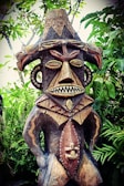 A wooden tribal sculpture with intricate carvings stands amidst lush green foliage. The figure features exaggerated facial features, including large eyes and a toothy mouth, and is adorned with decorative patterns. The sculpture's headpiece is adorned with bird motifs, adding to its elaborate design.