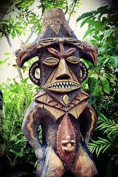 A wooden tribal sculpture with intricate carvings stands amidst lush green foliage. The figure features exaggerated facial features, including large eyes and a toothy mouth, and is adorned with decorative patterns. The sculpture's headpiece is adorned with bird motifs, adding to its elaborate design.