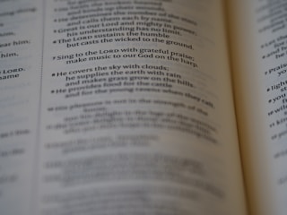 A close-up of an open book, focusing on a passage of religious text that advises singing to the LORD with grateful praise and making music to God on the harp. The text seems to be from a section of the Bible, displayed in crisp, readable font with slightly blurred pages in the background.