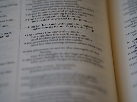 A close-up of an open book, focusing on a passage of religious text that advises singing to the LORD with grateful praise and making music to God on the harp. The text seems to be from a section of the Bible, displayed in crisp, readable font with slightly blurred pages in the background.