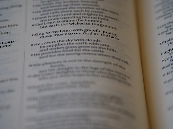 A close-up of an open book, focusing on a passage of religious text that advises singing to the LORD with grateful praise and making music to God on the harp. The text seems to be from a section of the Bible, displayed in crisp, readable font with slightly blurred pages in the background.