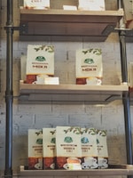 Coffee bags are arranged on a wooden shelf against a white brick wall. A variety of coffee types are displayed in branded packaging with cups and coffee illustrations.