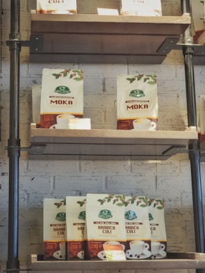 Coffee bags are arranged on a wooden shelf against a white brick wall. A variety of coffee types are displayed in branded packaging with cups and coffee illustrations.