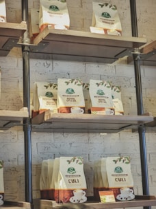 Bags of coffee are displayed on industrial-style metal and wood shelves against a white brick wall. The shelves are filled with various types of coffee bags including 'Robusta,' 'Royal Special,' and 'Culi.' Each bag has a logo and a small image of a cup of coffee on it.