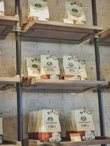 Bags of coffee are displayed on industrial-style metal and wood shelves against a white brick wall. The shelves are filled with various types of coffee bags including 'Robusta,' 'Royal Special,' and 'Culi.' Each bag has a logo and a small image of a cup of coffee on it.