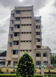 A multi-story building with a sign on top indicating 'Rajagiri College'. The structure has a modern design with multiple windows and a beige color. In the foreground, there is well-maintained greenery, including a large bush and a landscaped area. The sky appears overcast, suggesting a cloudy day.