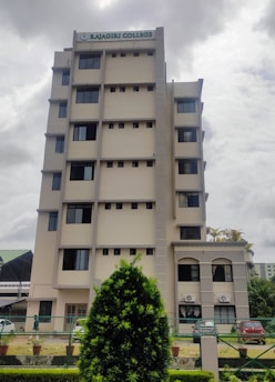 A multi-story building with a sign on top indicating 'Rajagiri College'. The structure has a modern design with multiple windows and a beige color. In the foreground, there is well-maintained greenery, including a large bush and a landscaped area. The sky appears overcast, suggesting a cloudy day.