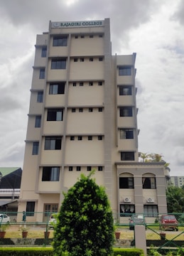 A multi-story building with a sign on top indicating 'Rajagiri College'. The structure has a modern design with multiple windows and a beige color. In the foreground, there is well-maintained greenery, including a large bush and a landscaped area. The sky appears overcast, suggesting a cloudy day.
