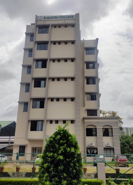 A multi-story building with a sign on top indicating 'Rajagiri College'. The structure has a modern design with multiple windows and a beige color. In the foreground, there is well-maintained greenery, including a large bush and a landscaped area. The sky appears overcast, suggesting a cloudy day.