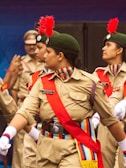 A group of young women in uniform performing a marching routine. They are wearing khaki uniforms with red sashes, green caps adorned with red feathers, and white gloves. Their uniforms display badges and colorful shoulder cords. The background shows other people in similar uniforms.