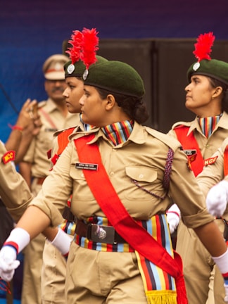 A group of young women in uniform performing a marching routine. They are wearing khaki uniforms with red sashes, green caps adorned with red feathers, and white gloves. Their uniforms display badges and colorful shoulder cords. The background shows other people in similar uniforms.