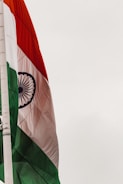 An Indian flag featuring horizontal tricolors of deep saffron, white, and India green with the blue Ashoka Chakra in the center. The flag is attached to a flagpole against a light sky.