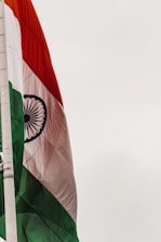 An Indian flag featuring horizontal tricolors of deep saffron, white, and India green with the blue Ashoka Chakra in the center. The flag is attached to a flagpole against a light sky.