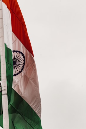 An Indian flag featuring horizontal tricolors of deep saffron, white, and India green with the blue Ashoka Chakra in the center. The flag is attached to a flagpole against a light sky.