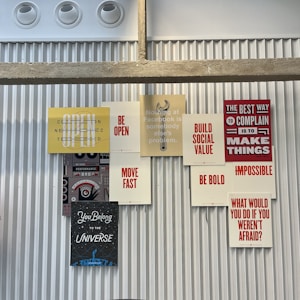 A collection of motivational and inspirational posters arranged on a corrugated metal wall. The posters feature phrases such as 'BE OPEN', 'MOVE FAST', 'BE BOLD', and 'BUILD SOCIAL VALUE' in bold typography. The display includes a stencil-style graphic of a wrench and other elements that convey a tech and innovation theme.