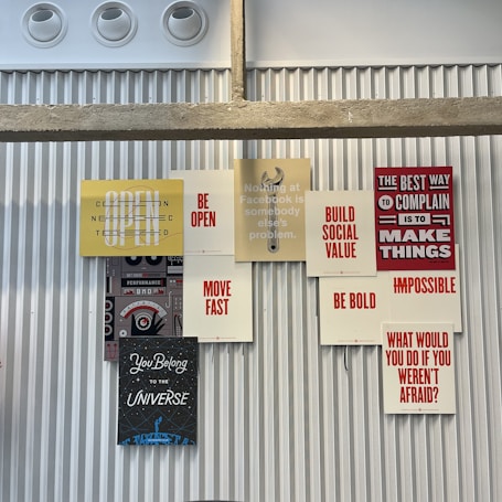 A collection of motivational and inspirational posters arranged on a corrugated metal wall. The posters feature phrases such as 'BE OPEN', 'MOVE FAST', 'BE BOLD', and 'BUILD SOCIAL VALUE' in bold typography. The display includes a stencil-style graphic of a wrench and other elements that convey a tech and innovation theme.