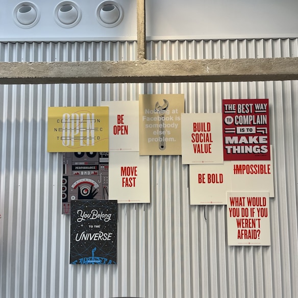 A collection of motivational and inspirational posters arranged on a corrugated metal wall. The posters feature phrases such as 'BE OPEN', 'MOVE FAST', 'BE BOLD', and 'BUILD SOCIAL VALUE' in bold typography. The display includes a stencil-style graphic of a wrench and other elements that convey a tech and innovation theme.