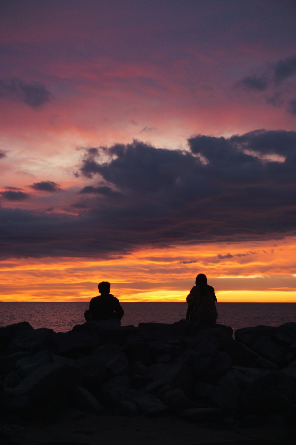 a couple of people sitting on a rock looking at the sunset