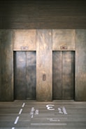Two elevator doors made of metal are set within a wall, each with a small display panel above. The floor in front of the elevators has text and directional arrows marked on it, using a light-colored stencil style.
