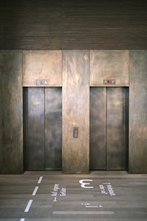 Two elevator doors made of metal are set within a wall, each with a small display panel above. The floor in front of the elevators has text and directional arrows marked on it, using a light-colored stencil style.
