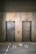 Two elevator doors made of metal are set within a wall, each with a small display panel above. The floor in front of the elevators has text and directional arrows marked on it, using a light-colored stencil style.