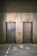 Two elevator doors made of metal are set within a wall, each with a small display panel above. The floor in front of the elevators has text and directional arrows marked on it, using a light-colored stencil style.