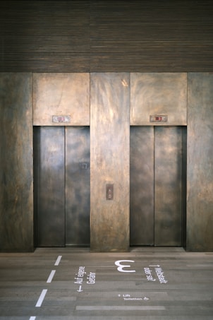 Two elevator doors made of metal are set within a wall, each with a small display panel above. The floor in front of the elevators has text and directional arrows marked on it, using a light-colored stencil style.