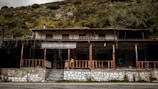 An old, abandoned building with a veranda and wooden railings. The structure appears weathered and is set against a backdrop of dense foliage and a hillside. The sign above the entrance contains Greek text.