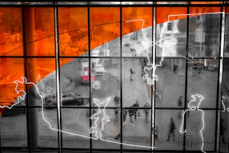 A large glass window with an abstract design featuring vibrant orange and black elements alongside white outlines. The window overlooks a street scene where people and a red vehicle can be seen blurred in the background.