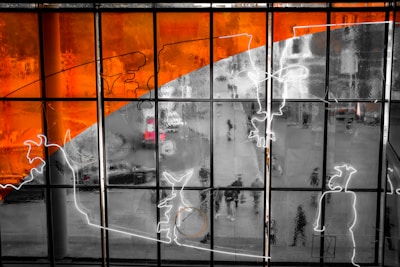 A large glass window with an abstract design featuring vibrant orange and black elements alongside white outlines. The window overlooks a street scene where people and a red vehicle can be seen blurred in the background.