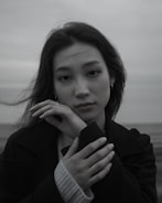 A black and white portrait of a person with long hair. The person looks directly at the camera with a gentle expression, hands softly resting under the chin. The background is an out-of-focus landscape.