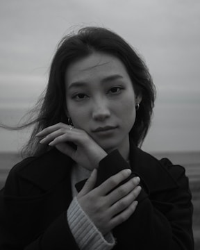 A black and white portrait of a person with long hair. The person looks directly at the camera with a gentle expression, hands softly resting under the chin. The background is an out-of-focus landscape.