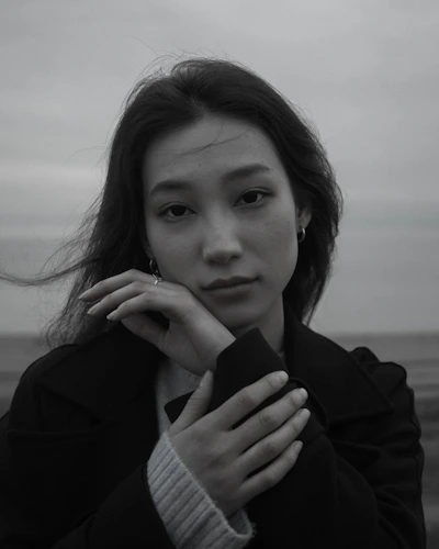 A black and white portrait of a person with long hair. The person looks directly at the camera with a gentle expression, hands softly resting under the chin. The background is an out-of-focus landscape.