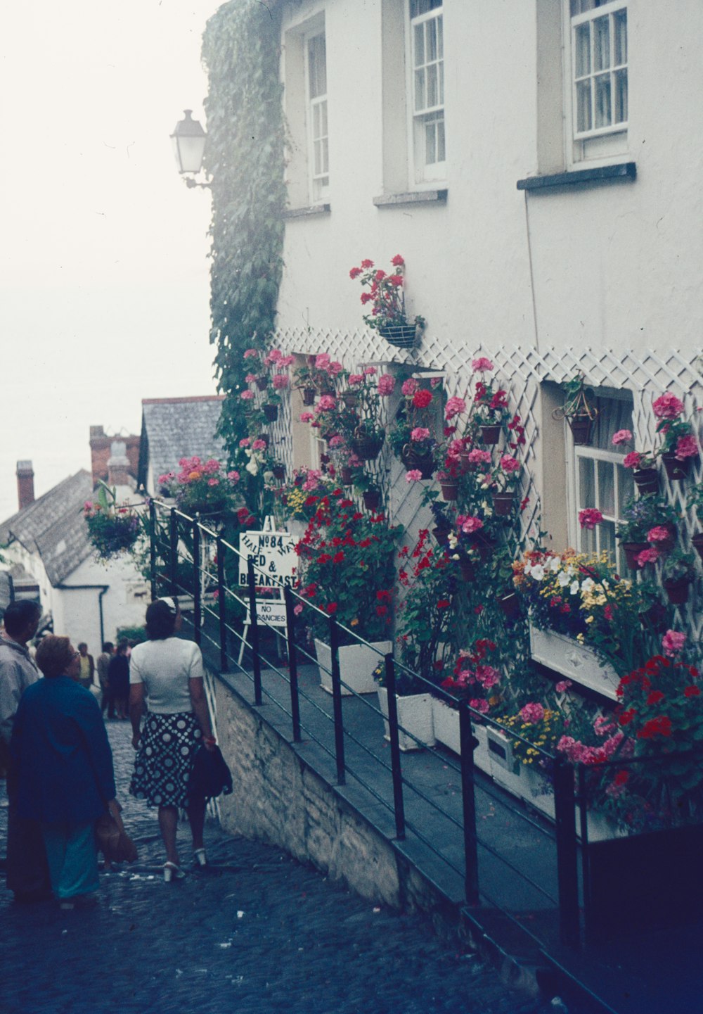 a group of people walking down a sidewalk next to a building with flowers