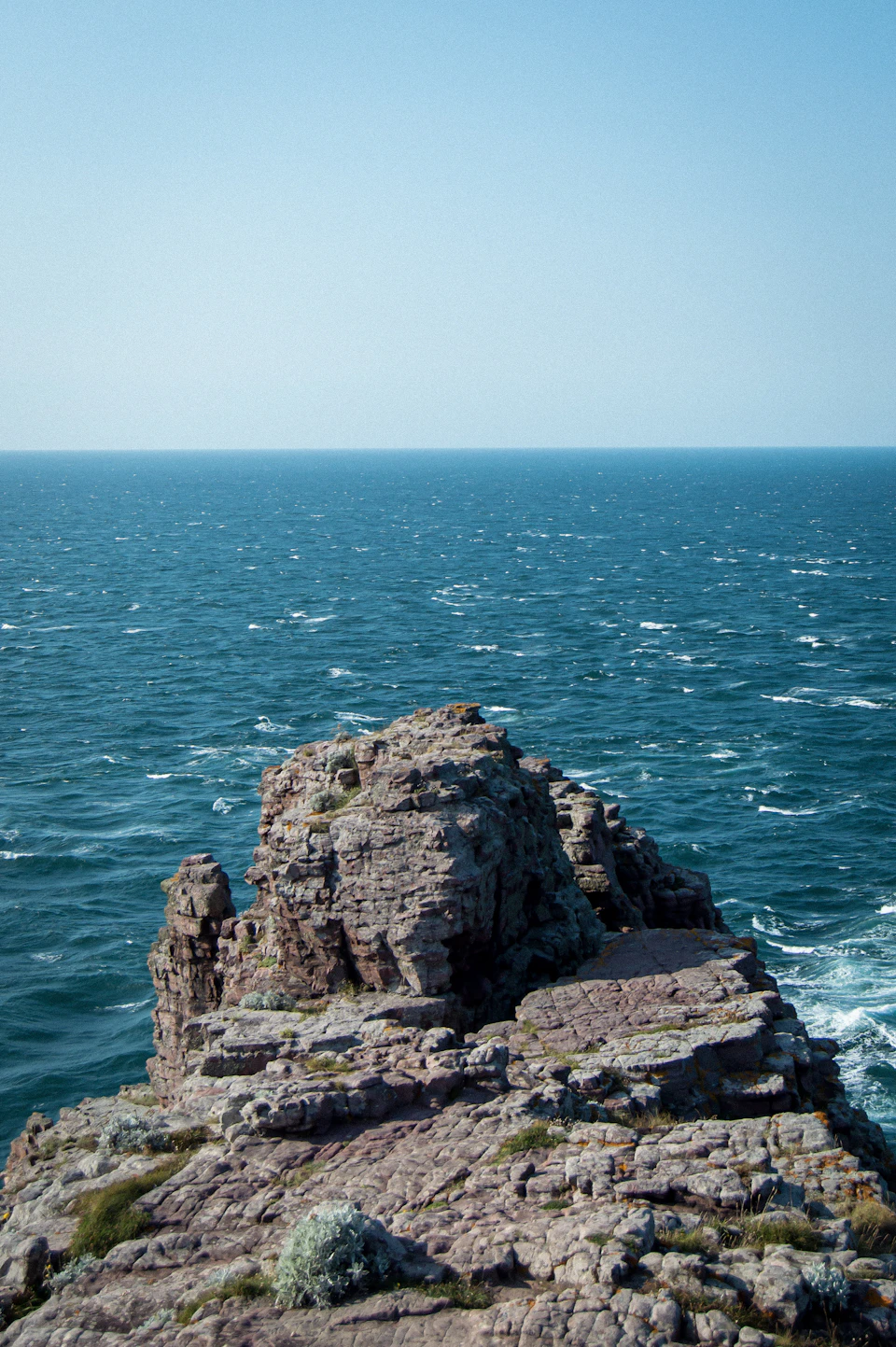 The image depicts a rocky cliff overlooking the ocean, with the vast expanse of blue water stretching out to meet the clear sky at the horizon. The perspective is from the top of the cliff, providing an unobstructed view of the serene landscape below. In the foreground, there are small plants and rocks scattered across the scene, adding texture and depth to the image.