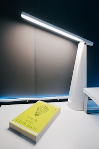 A white desk lamp is positioned on a light-colored desk, casting illumination on the wall behind it. Adjacent to the lamp is a yellow-cover book titled 'Think and Grow Rich' resting on the desk.