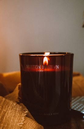A dark brown candle with a visible flame is placed on a wooden surface. The candle is in a glass holder, bearing the label of Molton Brown, a luxury brand. The warm, ambient lighting creates an inviting and cozy atmosphere, accentuated by a textured fabric draped underneath the candle.