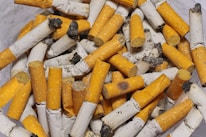 A pile of used cigarette butts with orange filters and white ash-covered paper, displaying signs of heavy use and ash debris.