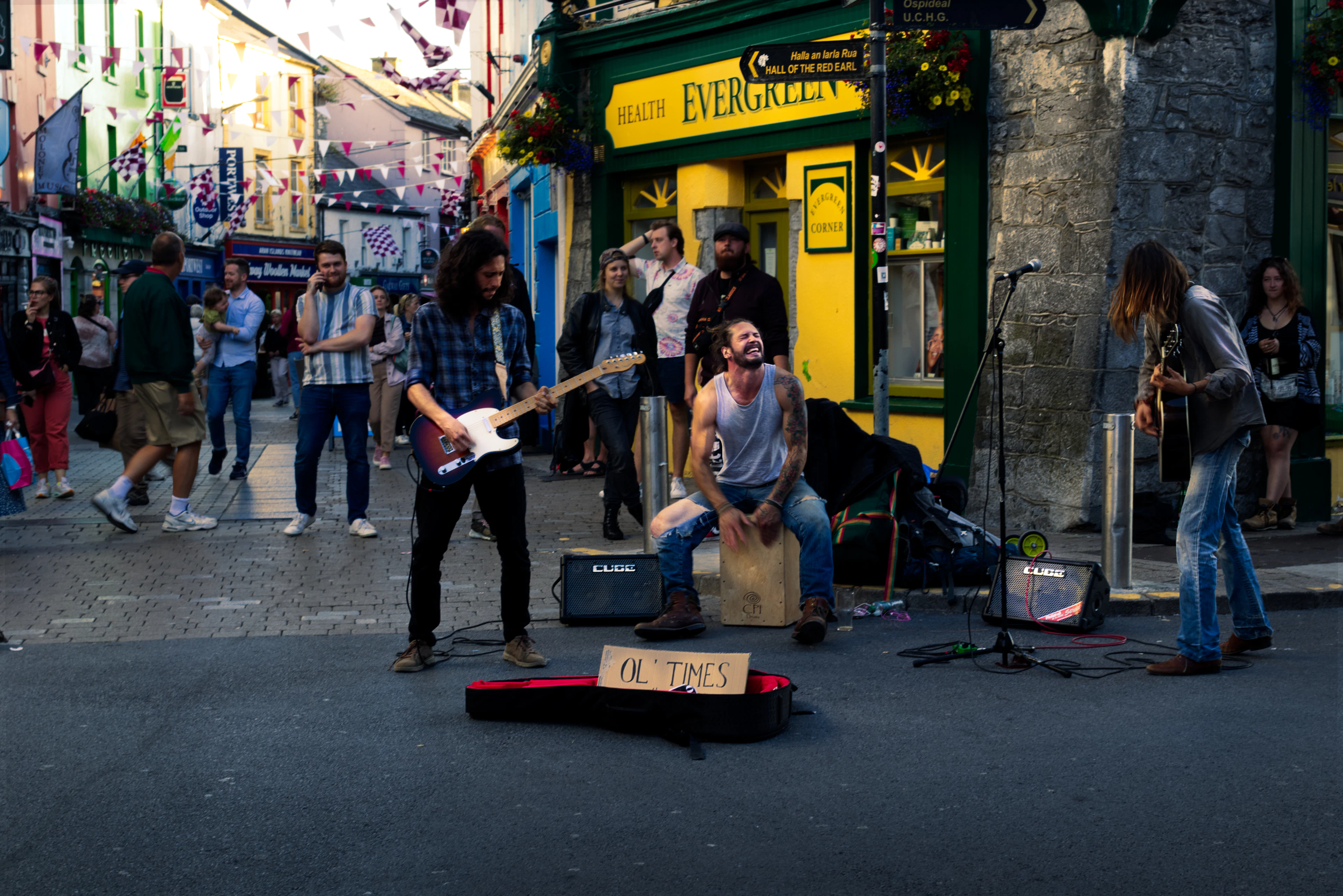 Three street musicians playing guitars and a cajon to a small crowd outside a bright green shopfront in Galway's Latin Quarter.