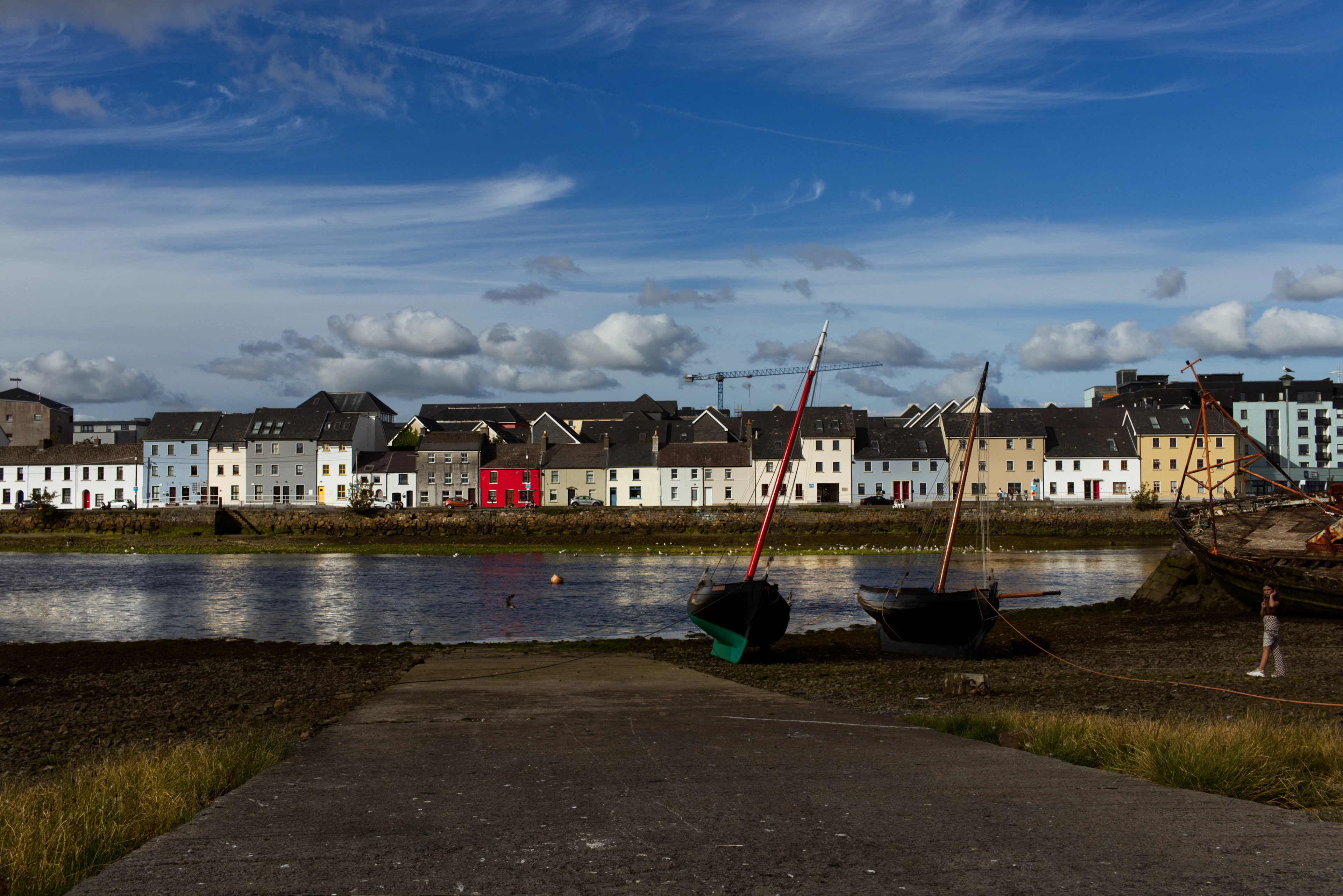 Two traditional Galway hookers with rust-red masts lying over on the mud at low tide in the Claddagh basin, the long white terrace of the quayside houses and a big Atlantic sky behind them.