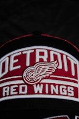 A close-up of a cap featuring the embroidered logo and text of the Detroit Red Wings, with a stylized winged wheel symbol prominently displayed. The words 'Detroit Red Wings' are written in bold white letters on a red background.