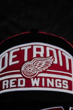 A close-up of a cap featuring the embroidered logo and text of the Detroit Red Wings, with a stylized winged wheel symbol prominently displayed. The words 'Detroit Red Wings' are written in bold white letters on a red background.