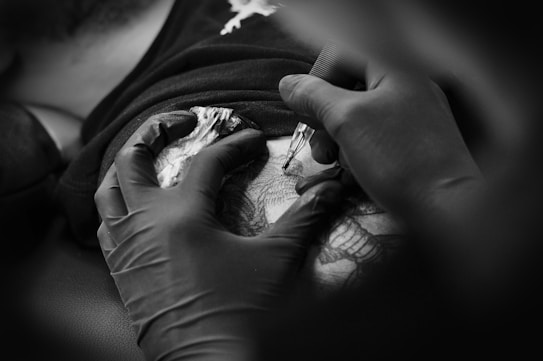 A person wearing black gloves is tattooing intricate designs on someone's skin. The scene appears focused and professional with attention to detail.